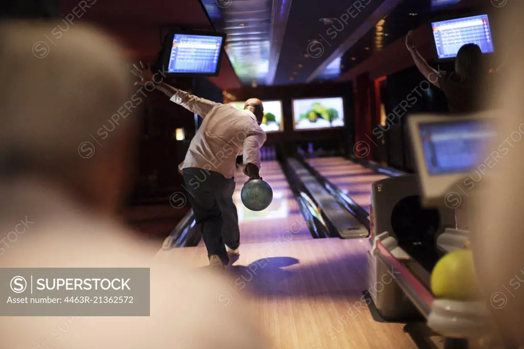 Mid adult man bowling a ball at a bowling alley watched on by friends.