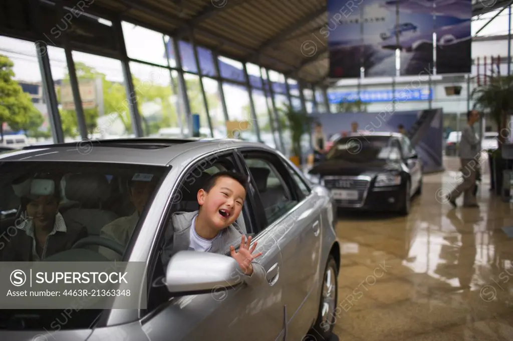 Portrait of a young boy waving out the window of a car he is sitting in with his parents inside a showroom.