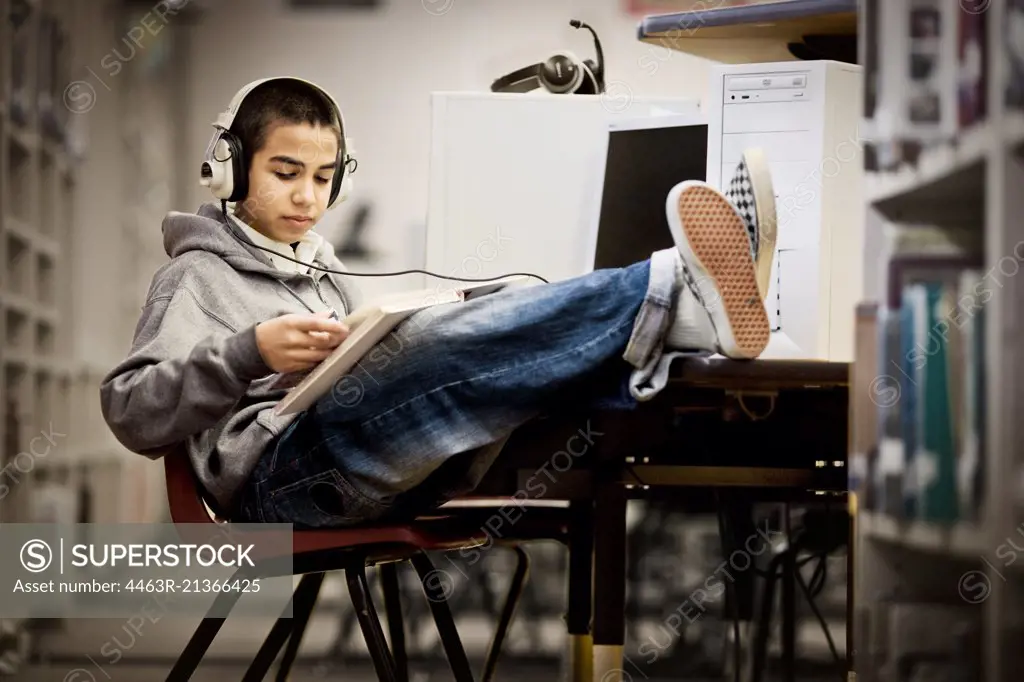 Teenage boy sitting with his feet up and reading a book with headphones on in a library.
