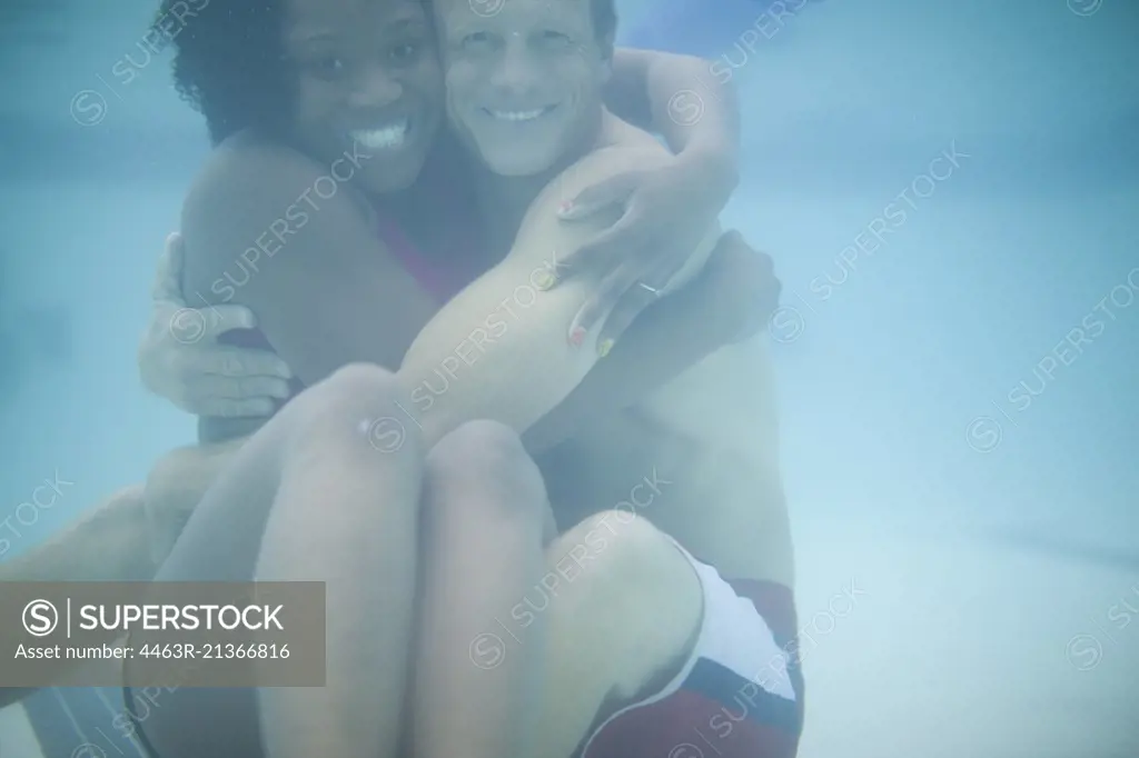 Portrait of a smiling young woman and mid-adult man hugging while sitting underwater in a swimming pool.