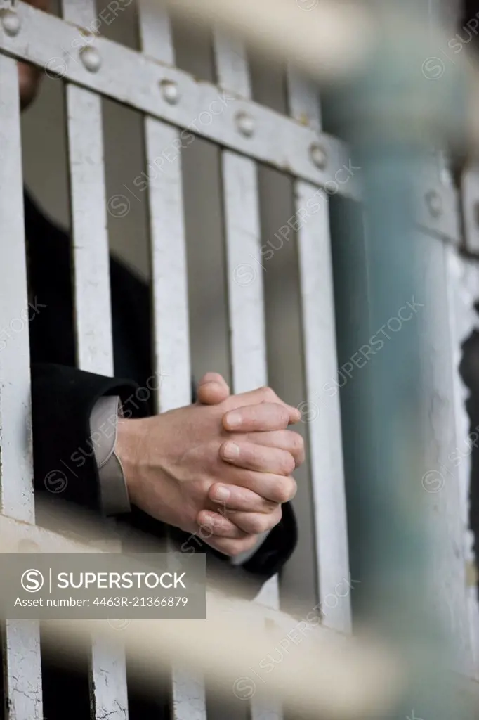 Hands of a young adult businessman through bars of a prison cell in a derelict building.