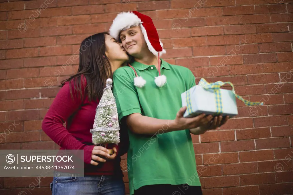 Teenage girl holding miniature Christmas tree kissing boyfriend on the cheek