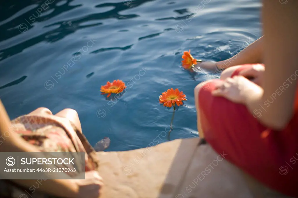 Flowers floating in a pool with woman dangling their feet in the water.