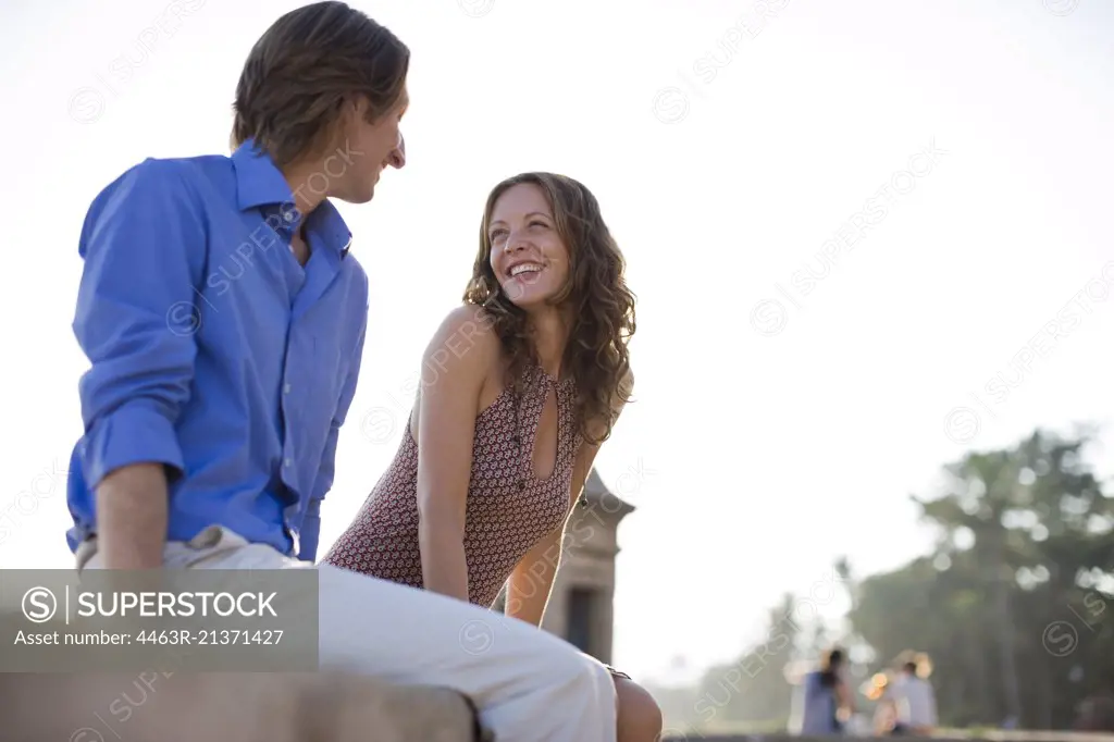 Mid-adult couple sitting on a stone wall looking at each other.