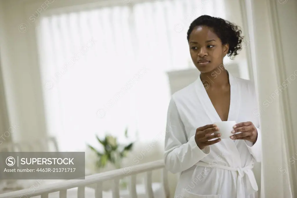 Young woman holding a cup while wearing a white robe inside her bedroom.