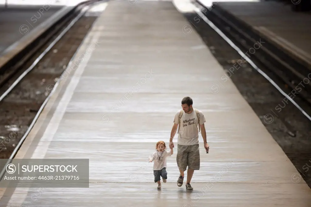 Father holds his toddler son's hand as they walk along a railway platform. 