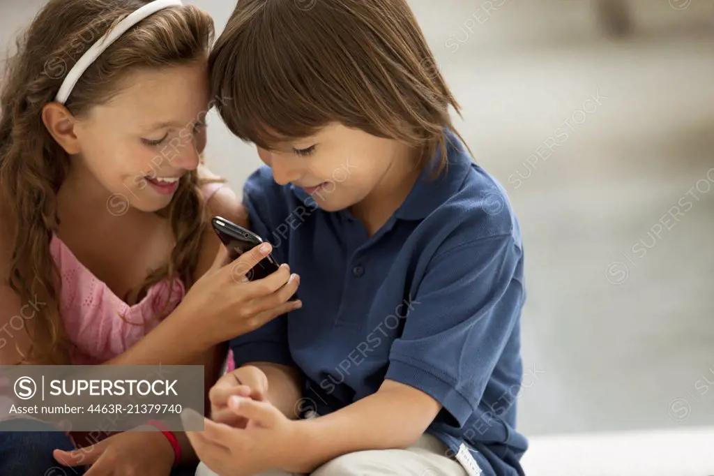 A young boy and girl smile mischievously as they look at a cellphone. 