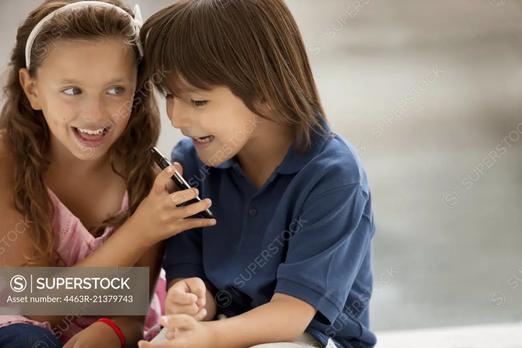 A young boy and girl smile mischievously as they look at a cellphone. 