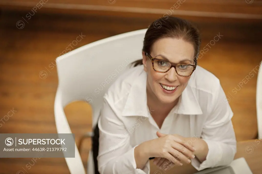 Businesswoman sitting at a desk with her hands clasped looks up and smiles as she poses for a portrait.