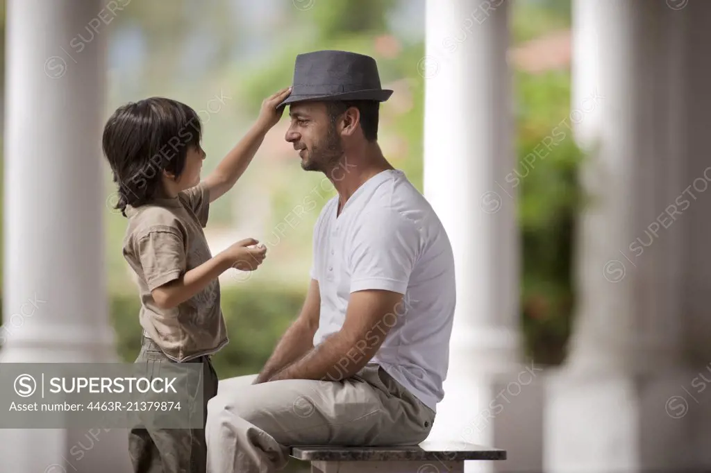 Thirtysomething man sits as his young son plays with his trilby hat.