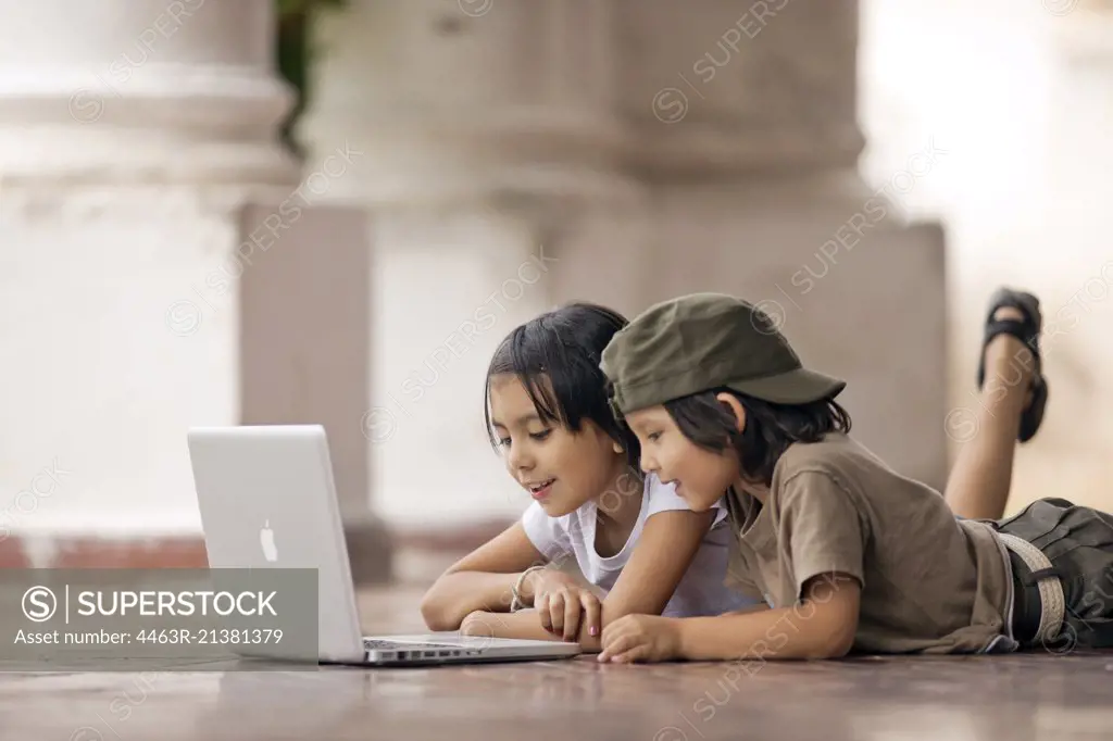 Brother and sister looking at a laptop on a tiled patio.