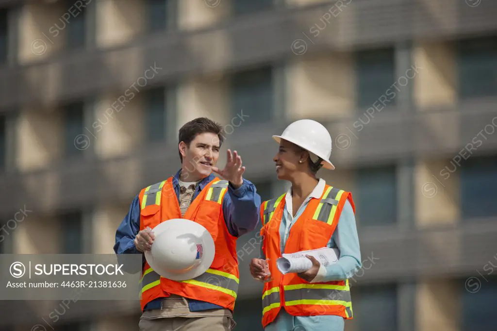 Two engineers in high visibility vests and hardhats discuss building plans on a construction site.