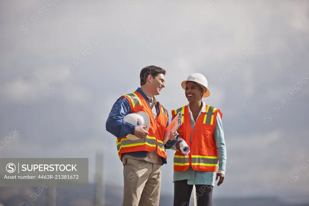 Two smiling engineers in high visibility vests and hardhats discuss building plans on a construction site.