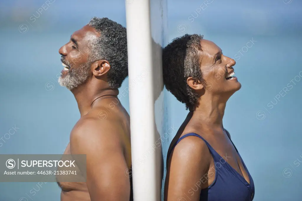 Laughing mature couple standing back to back on a beach.