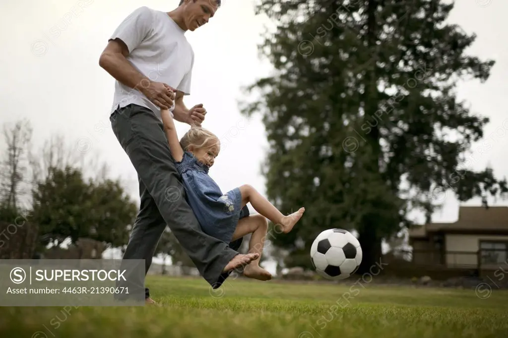 Smiling father teaches his little girl how to kick a soccer ball on the lawn.
