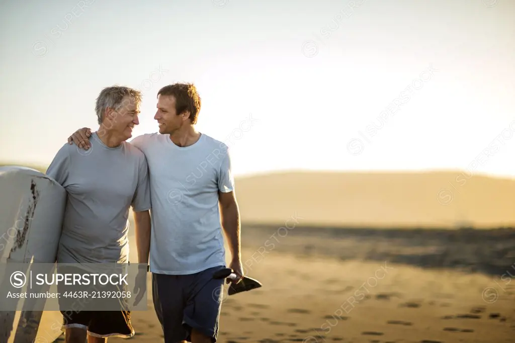Smiling father and middle aged son walking along a beach with their paddleboard.