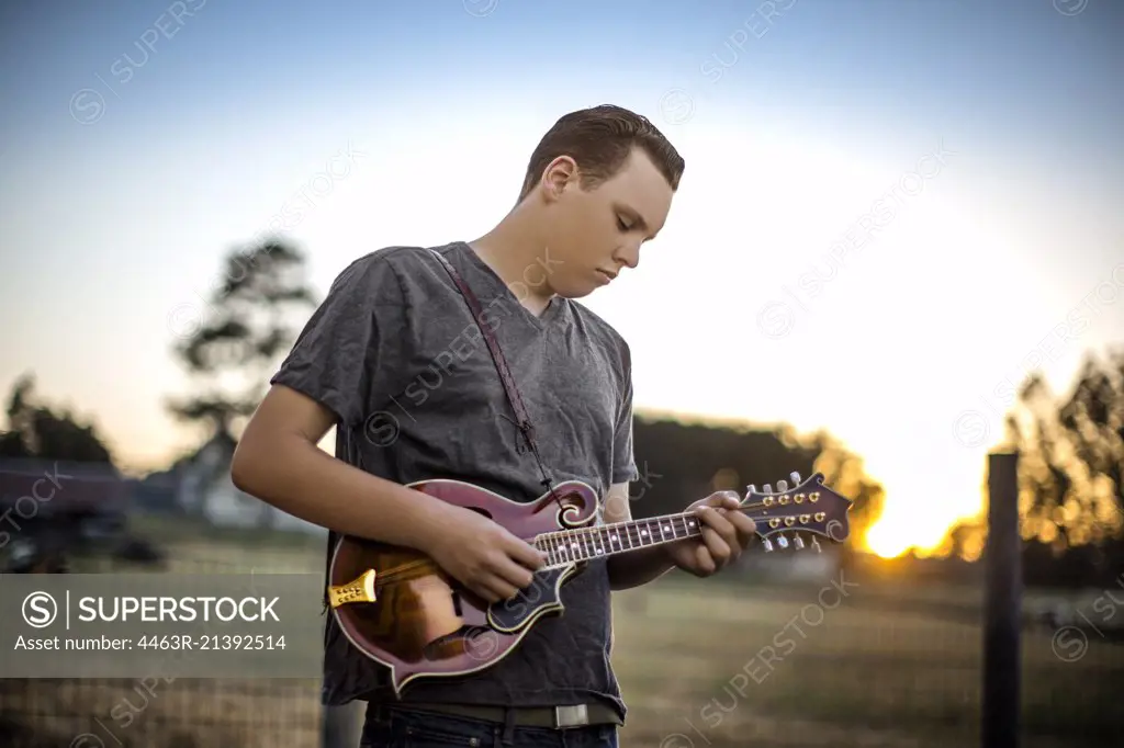 Contented teenage boy playing a mandolin in a rural field.