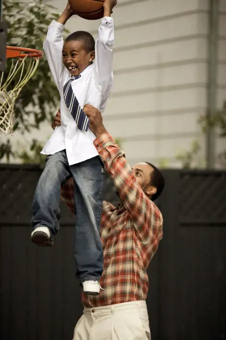Boy being held by his father while dunking a basketball.