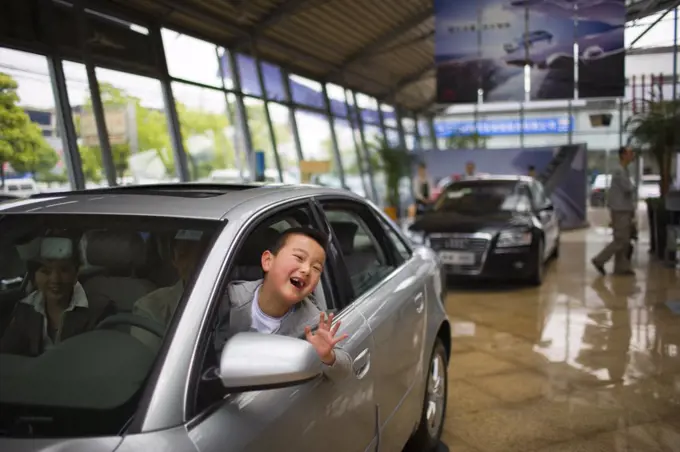 Portrait of a young boy waving out the window of a car he is sitting in with his parents inside a showroom.