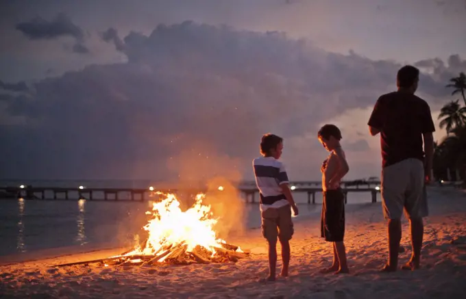 Man standing with to his two sons next to a camp fire on a sandy beach at dusk.