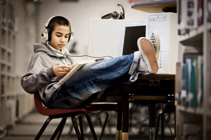Teenage boy sitting with his feet up and reading a book with headphones on in a library.