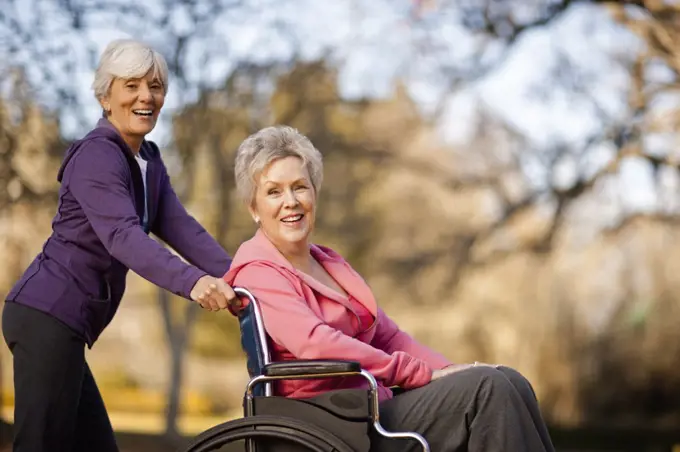 Portrait of a smiling senior woman being pushed through a park in a wheelchair by her friend.