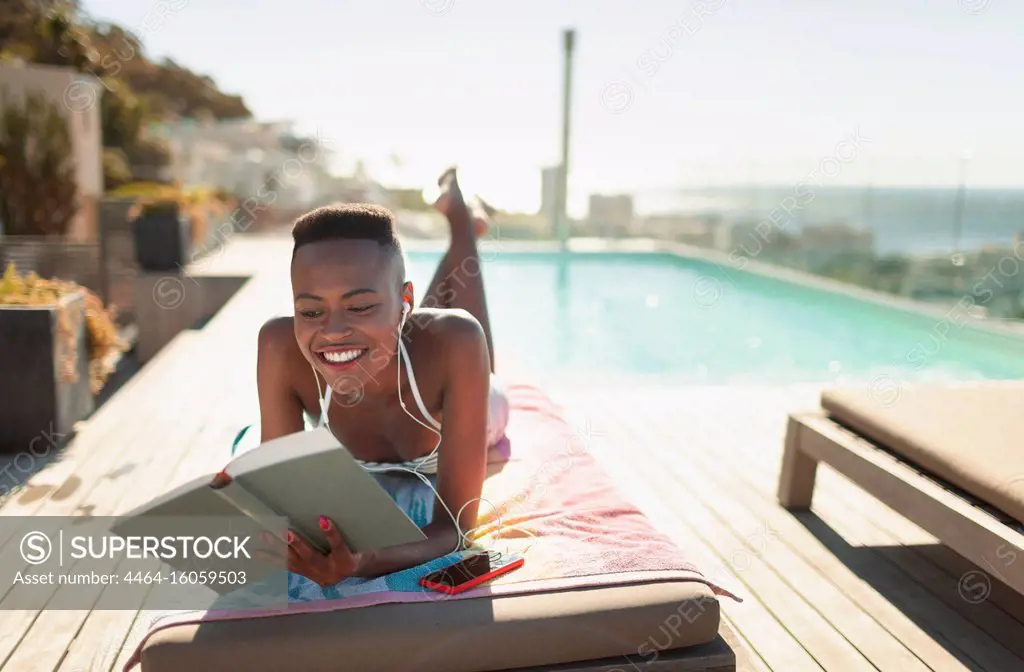 Smiling, carefree young woman reading book at sunny poolside