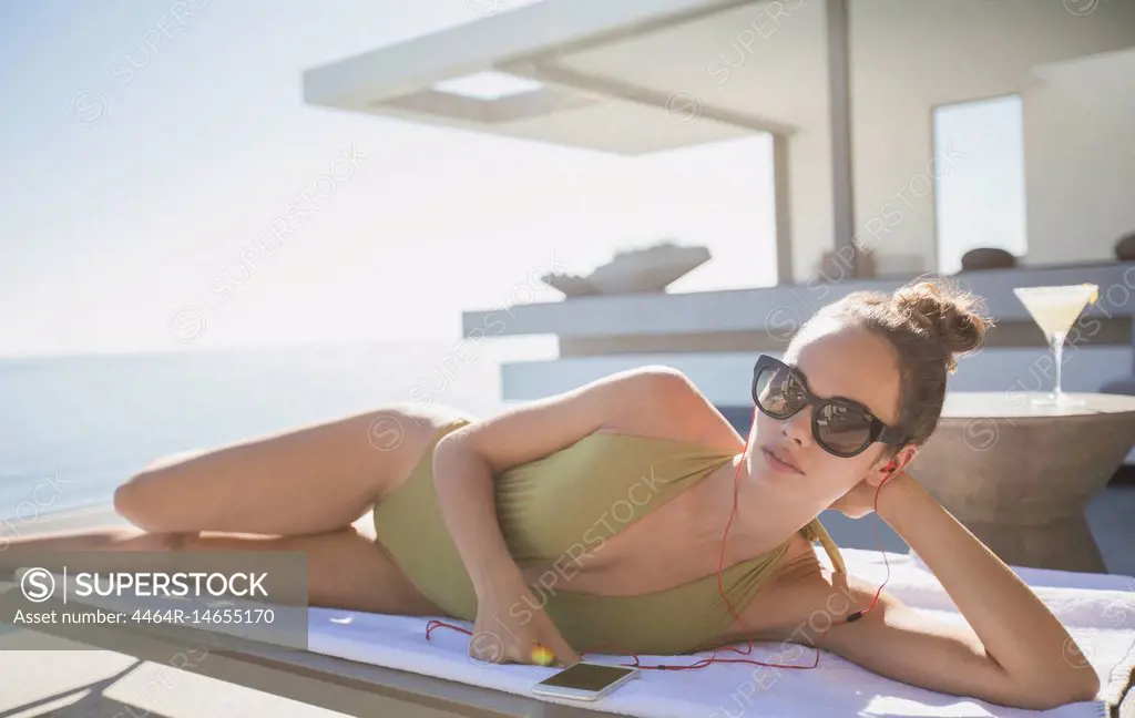 Portrait confident woman in bathing suit sunbathing on sunny luxury patio, listening to music with smart phone