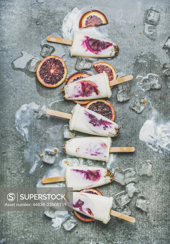 Healthy summer dessert. Blood orange, yogurt and granola popsicles on ice cubes over grey concrete background, top view, vertical composition. Clean eating, dieting, weight loss concept