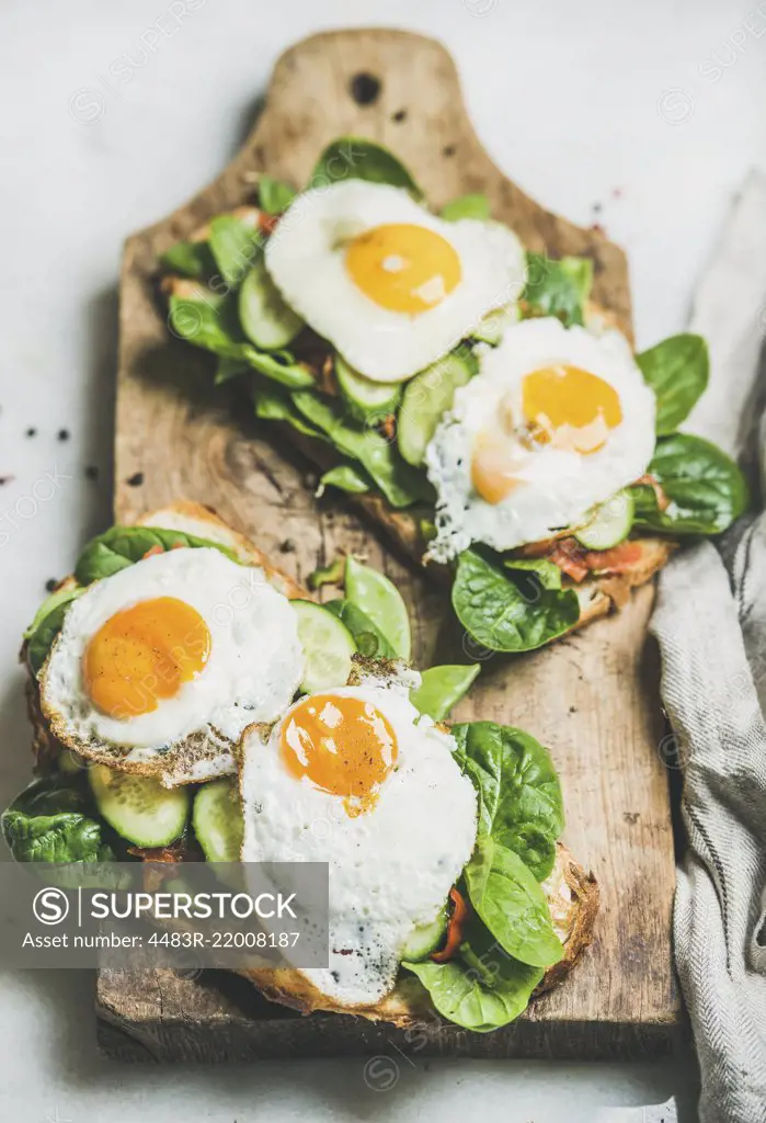 Healthy breakfast sandwiches. Bread toasts with fried eggs and fresh vegetables on wooden board over grey marble background, selective focus. Clean eating, healthy, weight loss, detox food concept