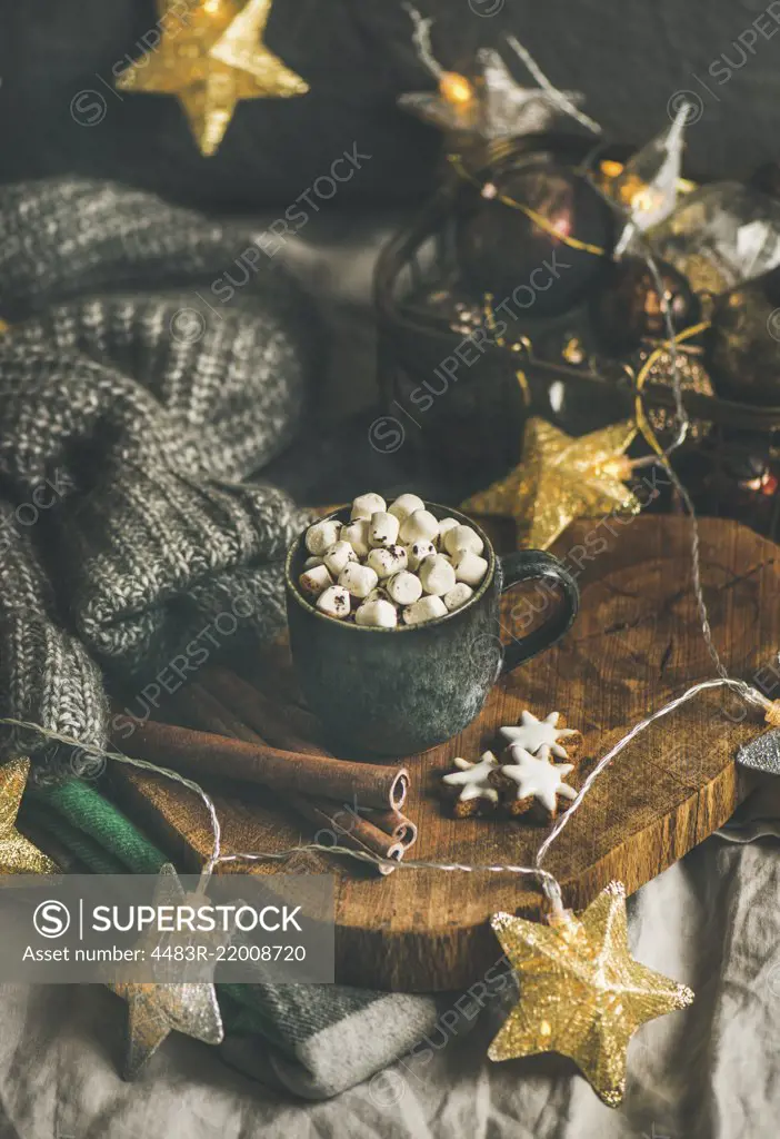 Christmas or New Year winter hot chocolate with marshmallows, gingerbread cookies and cinnamon sticks over wooden board served with holiday light garland and grey sweater, selective focus