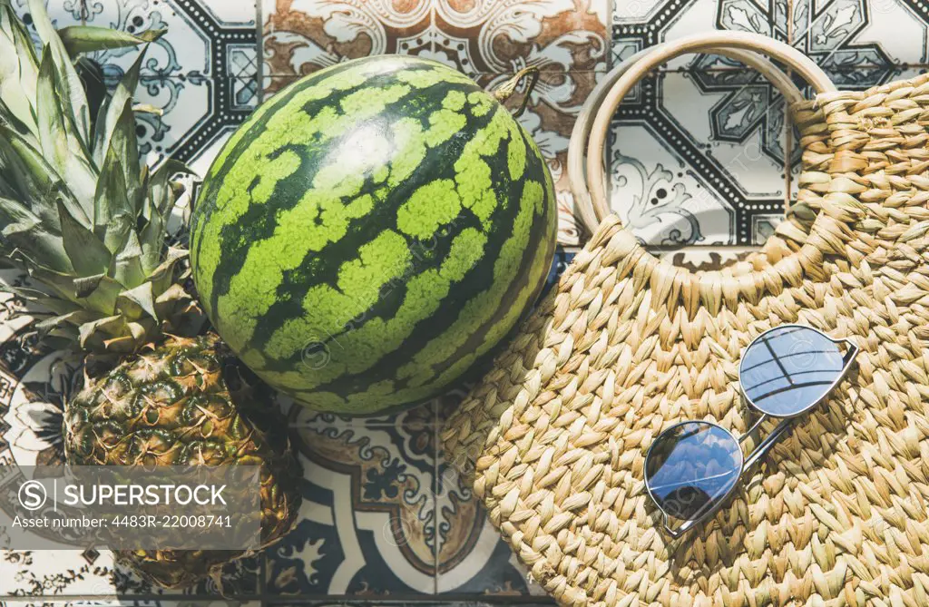 Summer lifestyle background. Flat-lay of summer fruit pineapple and watermelon, straw bag and sunglasses over colorful moroccan tile floor, top view