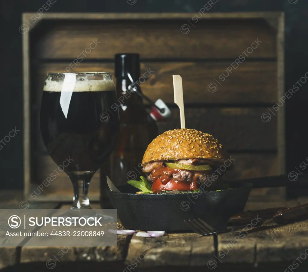 Homemade beef burger with crispy bacon, fresh vegetables and ketchup in black cast iron pan served with glass of dark beer on rustic wooden background, copy space, horizontal composition