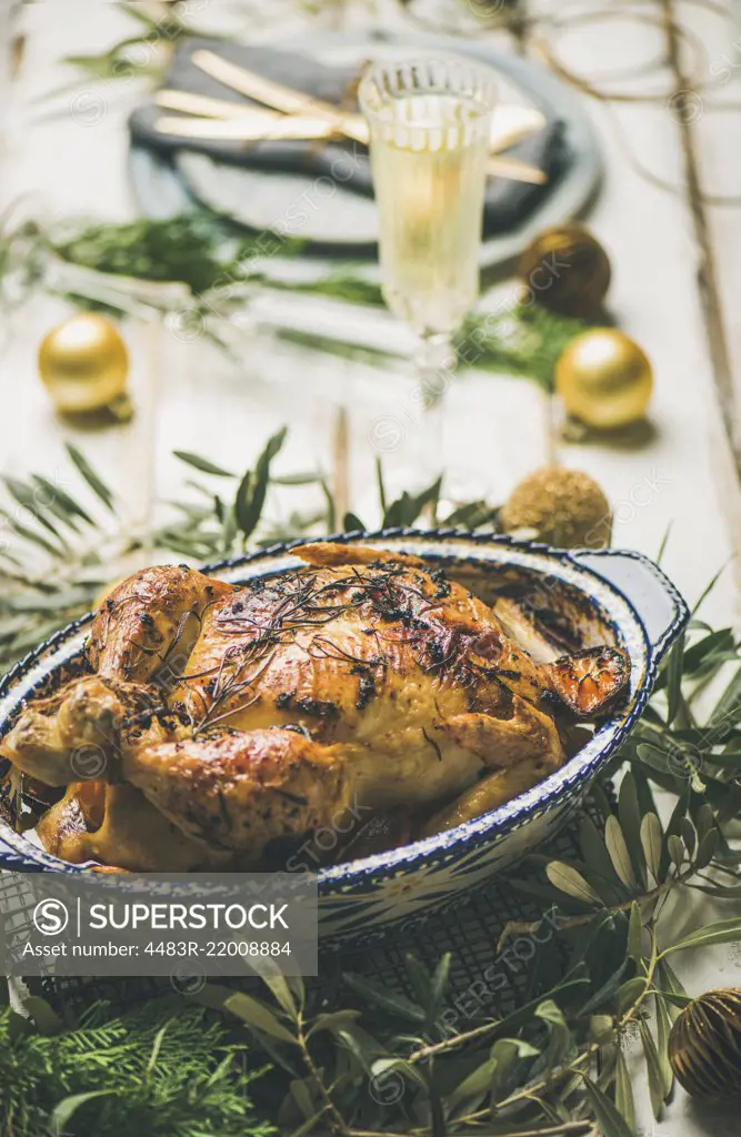 Christmas or New Year celebration table setting. Roast chicken, plates, silverware, glass and toy holiday decoration over rustic white wooden background, selective focus