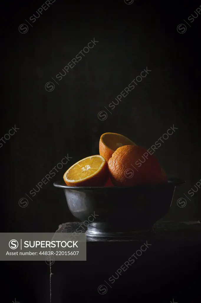 Whole and sliced oranges in vintage metal bowl over black table in the dark. Dark rustic style. With copy space at top