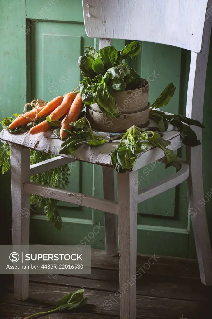 Bunch of fresh spinach and carrots on old white wooden chair with green wooden wall at background.