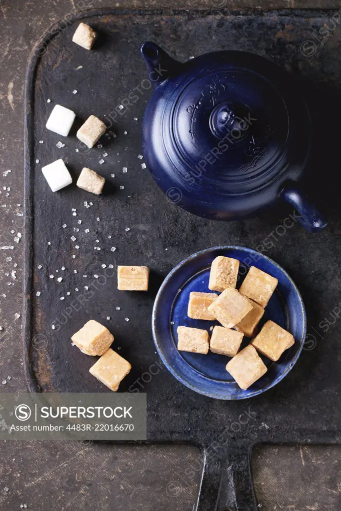 Blue ceramic plate with fudge candy ceramic teapot, served with sugar cubes over dark background. Top view