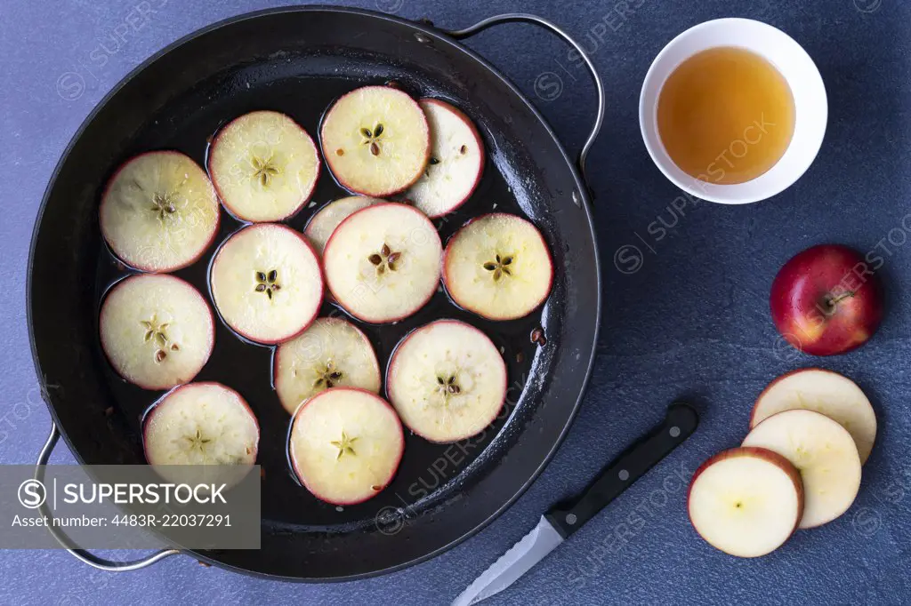 Sliced apples in a pan of honey syrup with a bowl of honey, a knife and a sliced apple.