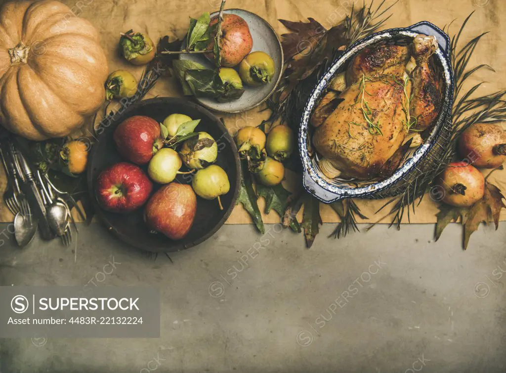 Thanksgiving dinner table. Flat-lay of roasted chicken or turkey, autumn fruit, pumpking, cutlery, leaves decoration over yellow linen table runner on grey concrete background, top view, copy space