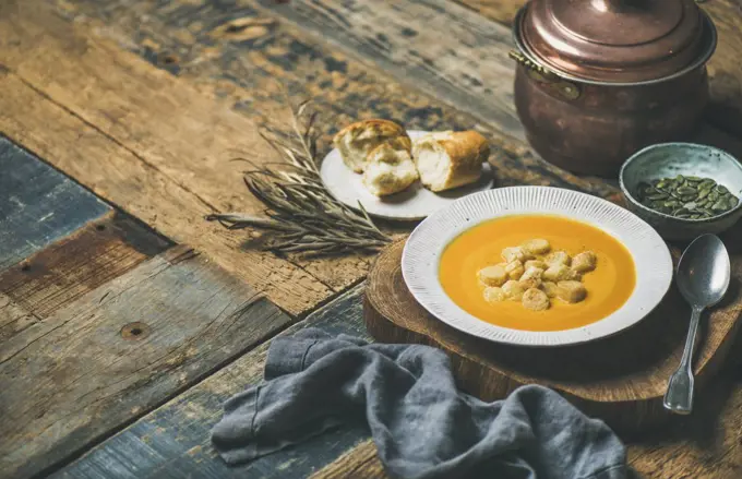 Fall warming pumpkin cream soup with croutons and seeds on board over rustic wooden background, copy space. Autumn vegetarian, vegan, healthy comfort food concept