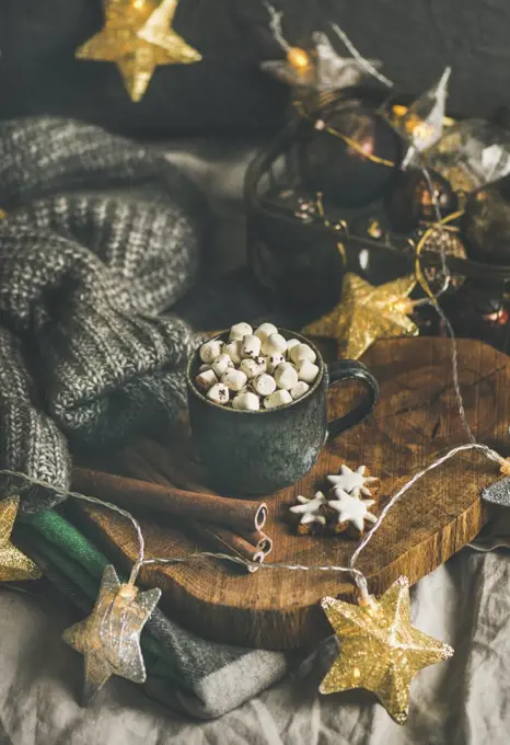 Christmas or New Year winter hot chocolate with marshmallows, gingerbread cookies and cinnamon sticks over wooden board served with holiday light garland and grey sweater, selective focus