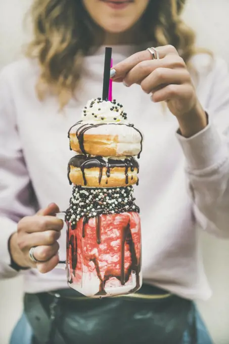 Young woman holding summer cold strawberry donut freakshake with whipped cream in mason jar in hands, selective focus