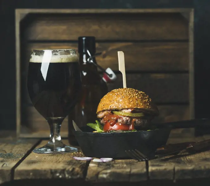 Homemade beef burger with crispy bacon, fresh vegetables and ketchup in black cast iron pan served with glass of dark beer on rustic wooden background, copy space, horizontal composition