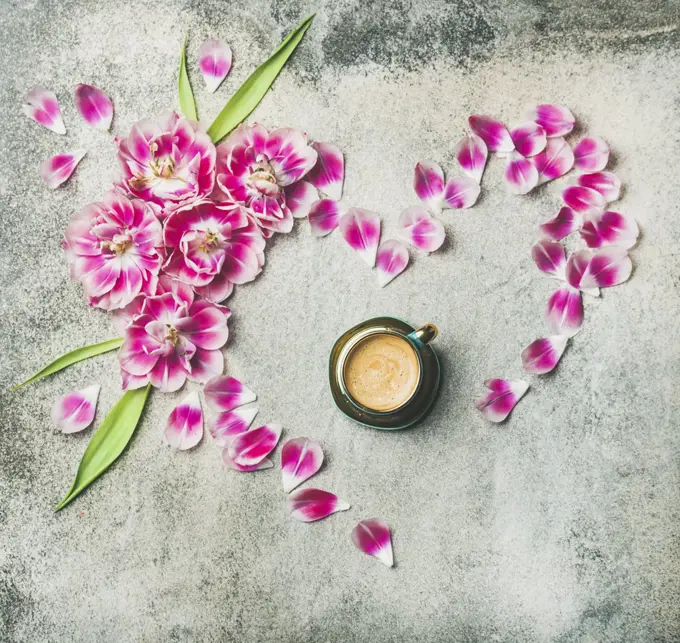 Spring morning concept. Flat-lay of cup of coffee surrounded with pink tulip flowers and petals in shape of heart over grey marble background, top view, copy space