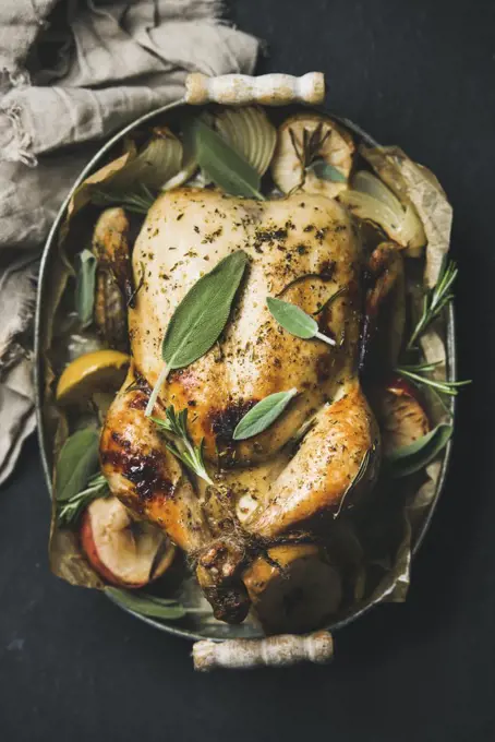 Oven roasted whole chicken with onion, apples and sage leaves in serving tray over dark stone background, top view, selective focus.