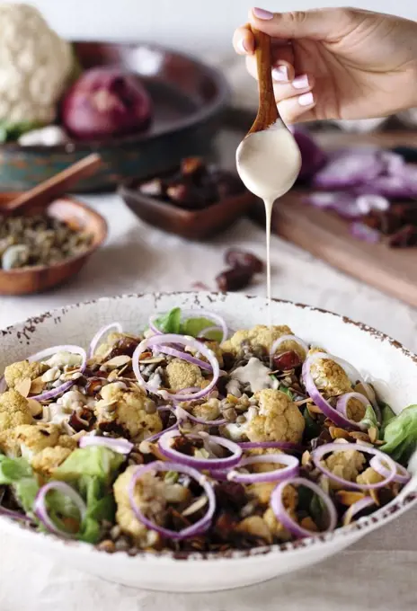 A woman is drizzling a bowl of roasted cauliflower salad with tahini dressing.