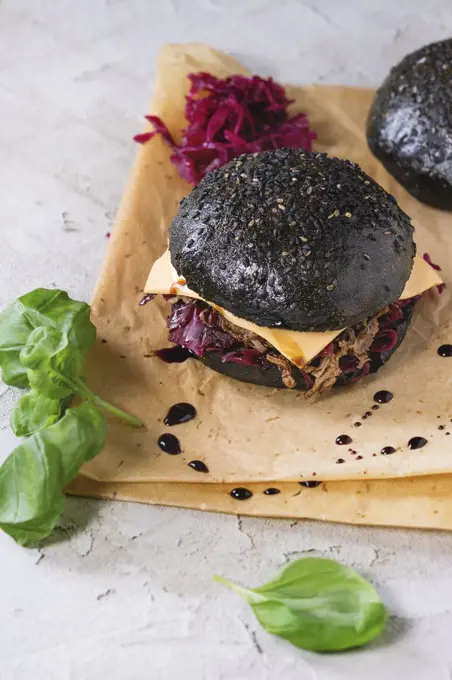 Black burger with beef stews, cheese, red cabbage and balsamic sauce served on baking paper with fresh basil over gray plastered surface