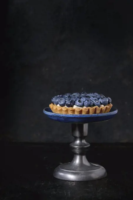 Lemon tartlet with fresh blueberries, served on vintage cake stand over black background.