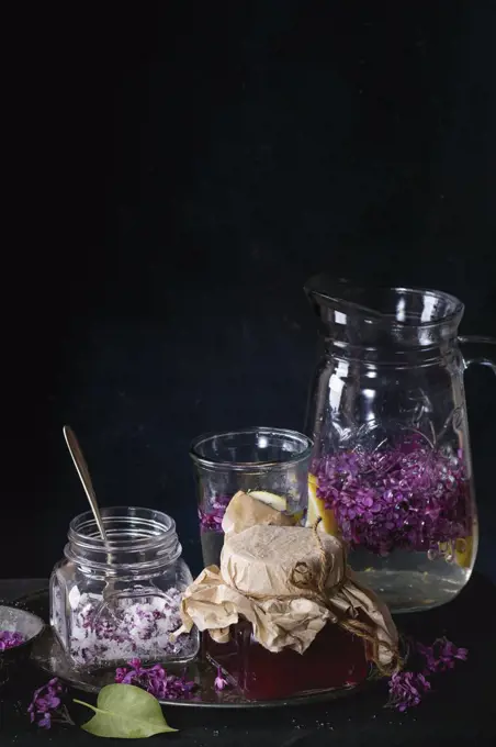 Glass jar of homemade lilac syrup, glass jar of sugared lilac flowers and glass pitcher of lilac lemonade water on black tablecloth over black. Dark rustic atmosphere.