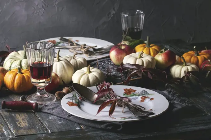Autumn holiday table decoration setting with decorative pumpkins, apples, red leaves, empty plate with vintage cutlery, red wine, candle over wooden table. Rustic style